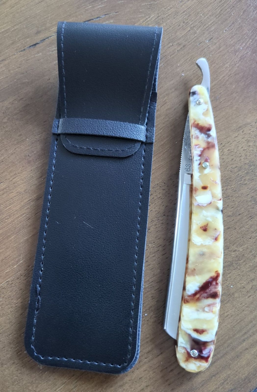 Black stitched razor case with a fold-over flap and tuck strap, beside a straight razor with marbled scales